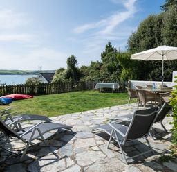 A garden with sun loungers a table with chairs and an umbrella by a fence overlooking water at Carn Du 1 in Rock