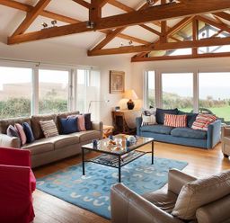 A living room with wooden beams sofas armchairs a coffee table and large windows overlooking greenery at Ossco Polzeath