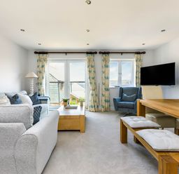 A living room with grey sofas a wooden coffee table a blue armchair a wooden dining table with cushioned benches and chairs a wall mounted tv and windows with floral curtains at Rock Lobster in Port Isaac