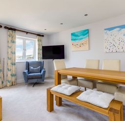 A dining area with a wooden table, white chairs, a wooden bench with cushions, a blue armchair, curtains with blue patterns, a wall-mounted TV, and two paintings on the wall at Rock Lobster in Port Isaac