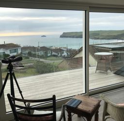 A room with a telescope, chairs, a small wooden table, and a glass door overlooking a deck and coastal houses at Atlantis Polzeath