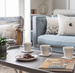 A coffee table with three cups on coasters a plate of biscuits a plant and a book in a living room with blue and patterned chairs at Albany in Port Isaac