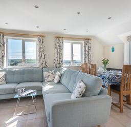 A living room with a light blue sectional sofa floral curtains and a dining table with wooden chairs at Polmorla in Polzeath