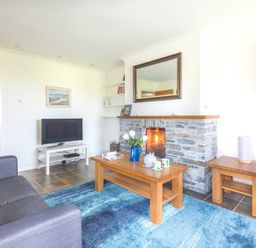 A living room with a grey sofa a wooden coffee table a stone fireplace a television on a white stand and a blue rug at Lundy Cottage in Rock