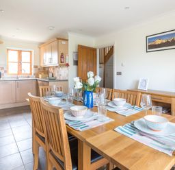 A dining table set with bowls glasses and cutlery next to a kitchen with wooden cabinets at Lundy Cottage in Rock