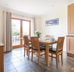 A dining room with a wooden table and six chairs set with dishes and glasses next to glass doors at Lundy Cottage in Rock