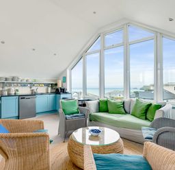 A living room with wicker chairs a sofa with green cushions a round table and a kitchen area with blue cabinets at The Point in Polzeath