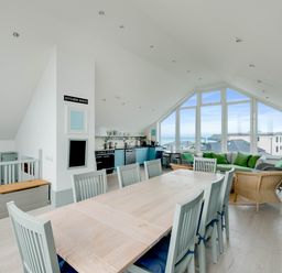 An open plan kitchen and dining area with a long wooden table and chairs a sitting area with green cushions and large windows showing a coastal view at The Point in Polzeath