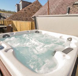 An outdoor hot tub with bubbling water on a wooden deck with stone buildings in the background at Cheviot Hills in Wooler