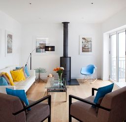 A living room with a white sofa and colorful pillows a glass coffee table a wood stove and a blue rocking chair at The Beach Hut in Polzeath