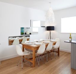 A dining area with a wooden table set for six and white chairs near a window at The Beach Hut in Polzeath
