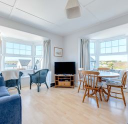 A living room with two blue armchairs by a window a wooden dining table with four chairs a TV on a wooden stand and two blue sofas at Pinewood Flat 5 in Polzeath