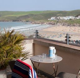 A balcony with a table and chairs overlooking a beach with people and houses on hills at Kellan in Polzeath