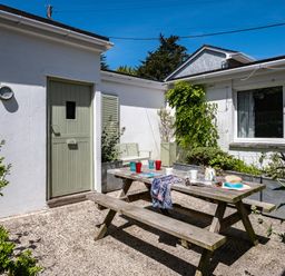 An outdoor patio area with a wooden picnic table set with cups plates and a hat beside a white building with a green door and window at St Michaels in Rock