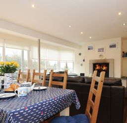 A dining table with cups and a vase of yellow flowers near windows and a living area with a fireplace at Pendaveli in Polzeath