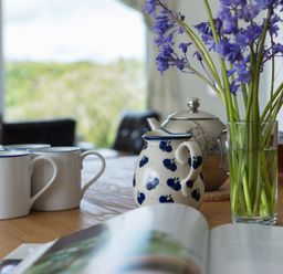 A wooden table with white mugs a patterned jug a metal teapot and a glass vase with purple flowers at Penny Liggan Rock