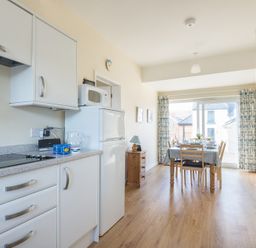 A kitchen area with white cabinets a dining table set with chairs and large window at The Holiday House (2 Atlantic Mews) in Polzeath