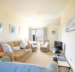 A living room with two wicker chairs a beige sofa a blue armchair a small television on a stand and framed pictures on the wall at The Holiday House 2 Atlantic Mews in Polzeath