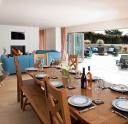 A dining area with a wooden table set with plates glasses and bottles next to a living room with blue sofas a fireplace and an open patio with outdoor seating at Downalong in Daymer Bay