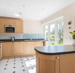 A kitchen with wooden cabinets a black countertop a stove a microwave a kettle flowers on a counter and glass doors overlooking a garden at Chy-An-Dowr in Rock