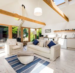 An open living room and kitchen area with wooden beams a sofa armchair and ottoman on a blue rug and built-in kitchen appliances at Greystones Studio in Daymer Bay