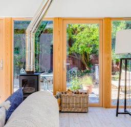 A living room with a wood stove and large windows showing a garden with chairs and plants at Greystones Studio in Daymer Bay