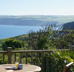 An outdoor wooden table with a cup, plate, and glass of orange juice with a wooden railing and coastal landscape at The Porthole in Port Isaac