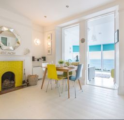 A dining area with a wooden table and colorful chairs next to a yellow-bricked fireplace and windows showing an ocean view at The View in Port Isaac