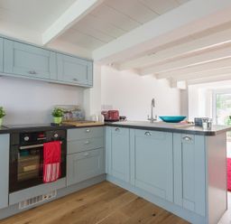 A kitchen with light blue cabinets black countertop and wooden floor adjacent to a dining area with a red rug at Beehive Cottage in Port Isaac