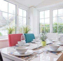 A dining table set with white plates bowls wine glasses and cutlery near windows with plants on the windowsill at Beehive Cottage in Port Isaac