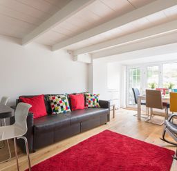 A living room with a black leather sofa with colorful cushions a red rug and a dining area with multicolored chairs at Beehive Cottage in Port Isaac