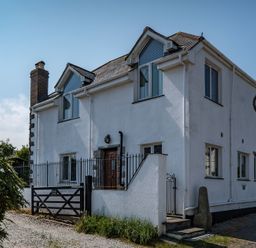 A white two-story house with a chimney and multiple windows surrounded by a black metal fence and gate at Fairholme in Port Isaac