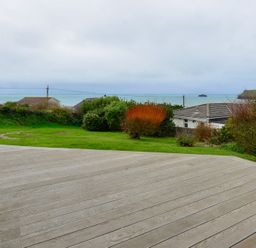 A garden with bushes and grass and a wooden deck overlooking houses and the sea at Sea Mist in Polzeath