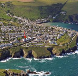 An aerial view of a coastal village with houses, roads, parking lots, green fields, cliffs, and ocean water at The Crows Nest in Port Isaac