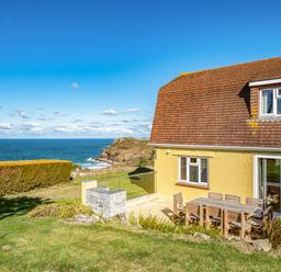 A yellow house with a brown roof next to a patio with wooden chairs and a table facing the sea at Trevan House Lundy in Polzeath