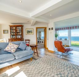 A living room with blue sofa wooden bookcase chairs and large windows showing ocean view at Trevan House Lundy in Polzeath