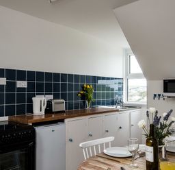 A kitchen with blue tiled backsplash a kettle toaster flowers on the counter a microwave and a dining table with plates glasses and flowers at Pinewood 14 in Polzeath