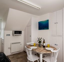 A kitchen dining area with a round table set for four, white chairs, a microwave on the wall, cupboards, and a window at Pinewood 14 in Polzeath