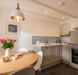 A kitchen with grey cabinets a wooden round dining table with flowers and plates at Kipper Cottage in Port Isaac