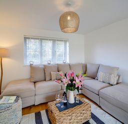 A living room with a beige sectional sofa a woven coffee table with flowers and wine glasses and a floor lamp near the window at Elmwood in Port Isaac