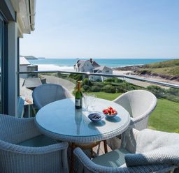 A balcony with a glass table and wicker chairs overlooking the ocean and a grassy area at Egloskerry in New Polzeath
