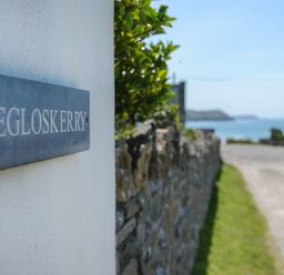 A sign reading Egloskerry on a white wall with a stone wall and a coastal road leading to the sea at Egloskerry in New Polzeath