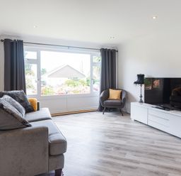 A living room with a gray sofa and pillows a gray armchair with a yellow cushion a large window with dark curtains a white TV stand a flat screen TV and a small lamp with a black shade at Greenaway 4 Rockhaven Gardens Rock