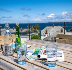 A wooden table with glasses of water a water bottle a green bottle and magazines outdoors with a sea view at Seahaven in Port Isaac