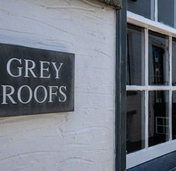 A black sign with white letters reading grey roofs on a white textured wall next to a window with white panes at Grey Roofs in Port Isaac