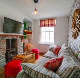 A living room with a striped sofa and red cushions a wooden chest coffee table a brick fireplace with a mounted TV and a window with striped blinds at Grey Roofs in Port Isaac