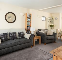 A living room with a gray sofa, armchair, wooden bookshelf, side table with lamp, and dining table at Sea Glimpses in Port Isaac