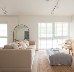A living room with a beige sofa with cushions a large mirror a window with blinds an ottoman and a wooden cabinet at Westward 8 in Polzeath