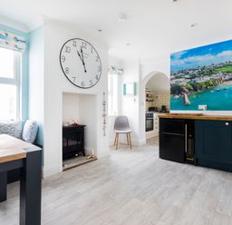 A dining area with a wooden table and bench seating beside windows and a kitchen with navy cabinets and a large coastal wall mural at Number 28 in Port Isaac