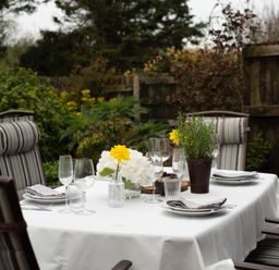 An outdoor dining table with a white tablecloth set with plates glasses napkins and floral centerpieces surrounded by striped cushioned chairs in a garden